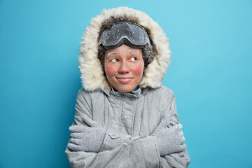 Frozen woman embraces herself trembles from cold covered with hoarfrost looks gladfully wears snowboard goggles winter jacket and gloves smiles pleasantly isolated over blue studio background