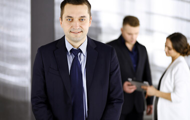 Portrait of a self-confident middle aged businessman in a blue suit, standing in a modern office with his colleagues at the background. Concept of business success