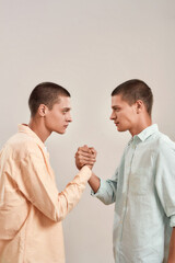 Vertical shot of young caucasian twin brothers looking at each other, holding hands while standing face to face isolated over beige background
