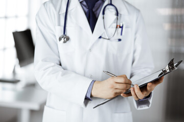 Unknown male doctor standing and working with clipboard of medication history records in clinic at his working place, closeup. Perfect medical service in hospital. Medicine and healthcare concept