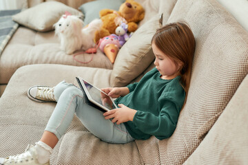 A little girl sitting on a sofa playing games on a tablet with her shoes on