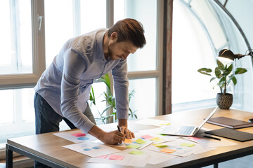 Thoughtful serious young businessman employee standing at desk focused on paperwork, making changes correcting mistakes in marketing plan by hand, analyzing client customer demands using infographics