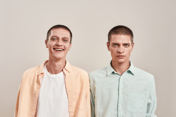 Portrait of two young caucasian men, twin brothers looking at camera with different face expressions while posing together isolated over beige background - obrazy, fototapety, plakaty