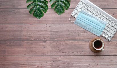 Wood table with keyboard, face mask, coffee and green plants.
