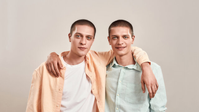 Portrait of two happy caucasian men, young twin brothers hugging and smiling at camera while posing together isolated over beige background