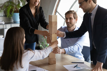 Smiling friendly diverse young staff engaged in building tower of wooden bricks as part of...