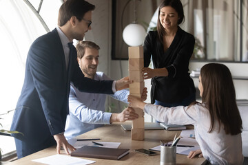 Happy excited multiethnic business team leaded by professional mentor coach participating in teambuilding activity playing educational game building tall column from cubes supporting helping coworkers