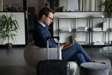 Wasting no time. Focused young businessman corporate worker being on assignment or business trip sitting in cozy office on arrival working on laptop pc online expecting for appointment negotiations