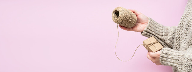 Women's hands hold a gift and a coil of rope on a pink background, close-up, copy space. Handmade...