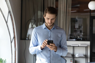 Focused young man banker consultant standing in office area concentrated on cellphone screen making answering call, texting message, giving distant consultation to client online, sending urgent email