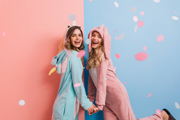 Dreamy girl in rabbit kigurumi holding hands with friend. Ecstatic ladies in cute pajamas dancing and smiling.