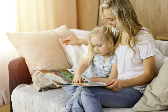 Happy family. Blonde young mother reading a book to her cute daughter while sitting at wooden floor in sunny room. Motherhood concept