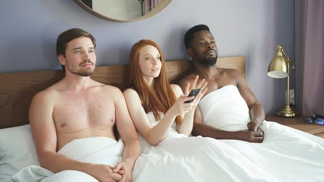 Diverse trio watches TV in the bedroom. Concept of free relationships. Red-haired woman sitting with two men in bed