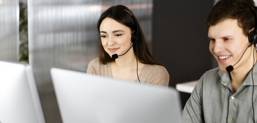 Young friendly man in headsets is talking to the client, while sitting at the desk with a female colleague in an office. Call center operators at work