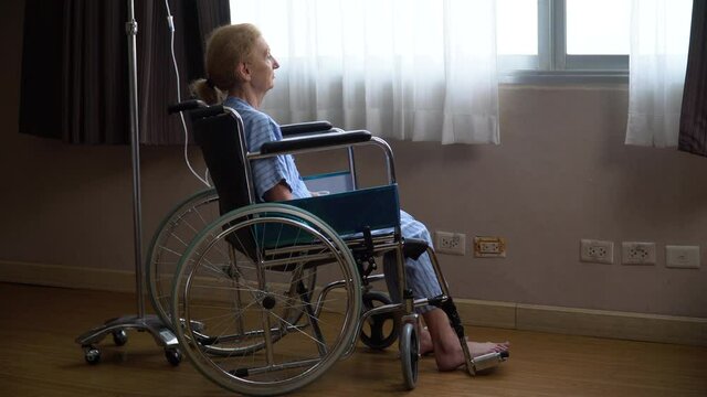 senior woman patient sit on wheelchair alone and looking out of window trying to stand up but failed at hospital ward. Lonely old female struggle with rheumatism in rehab Nursing home.