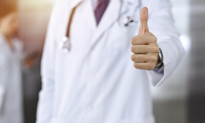 Unknown man-doctor standing straight and showing Ok sign with thumb up in modern clinic. Female colleague is in a hurry at the background of physician. Medicine concept