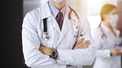 Unknown male doctor in protective mask standing with arms crossed in sunny clinic. Medicine concept...