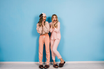 Full length view of positive girls in pajamas smiling at camera. Studio shot of pretty ladies in sleepwear standing on blue background.