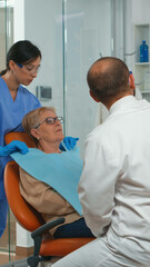 Stomatologist pointing on digital screen explaining x-ray to elderly woman. Doctor and nurse working together in modern stomatological clinic, examining, showing radiography of teeth on monitor