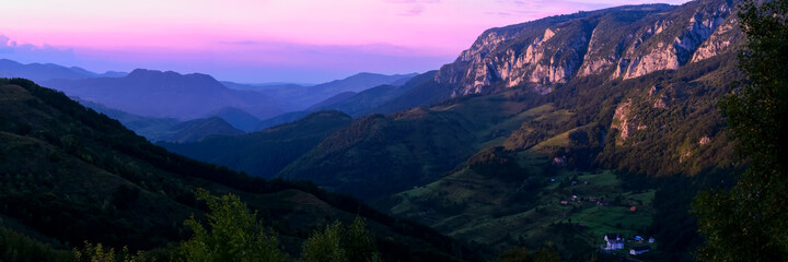 Idyllic panoramic landscape at sunset with purple sky during summer above the Apuseni mountains in the Carpathian arch with an isolated village in the foreground and mountains layers in the distance.