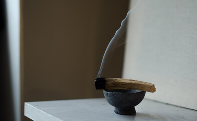 An image of a burning palo santo smudge stick in a stone bowl on a white ledge.