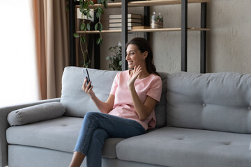 Fototapeta premium Relaxed young happy arabic indian mixed race woman sitting on sofa, involved in video call pleasant conversation using modern technology cellphone application, looking at web camera at home.