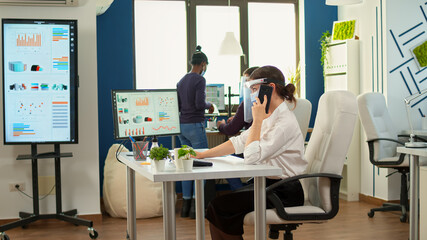 Freelancer with protection mask talking on mobile phone with partner, sitting at desk in office room during pandemic. Multiethnic coworkers working respecting social distance in financial company