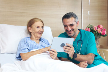 Doctor or assistance staff talking with an elder patient woman while using smart tablet and sitting on bed in the hospital