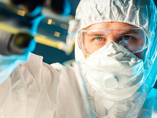 Man in a biological protection suit close-up. He is holding test tube in front of screen. Biologist in mask and protective coveralls. Concept - work with hazardous biological substances. Biology work