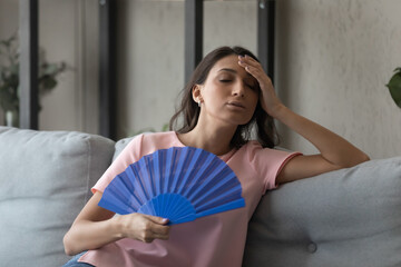 Overheated displeased millennial arabic indian mixed race woman using paper fan, suffering from hot summer weather or high temperature at home without air conditioning system, feeling unwell.