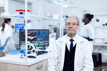 Naklejka na ściany i meble Senior man with virus on computer display looking at camera with protective glasses. Elderly scientist wearing lab coat working to develop a new medical vacine with african assistant in the background