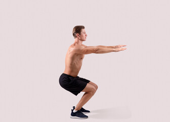 Obraz premium Side view of strong young guy doing squats on light studio background, full length portrait