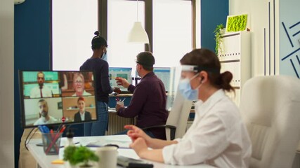 Businesswoman with face mask in office talking on video call with remote team during coronavirus epidemic. Manager having online conference while colleagues working respecting social distance - Powered by Adobe