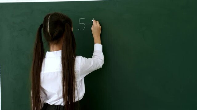 Little schoolgirl writing on chalkboard in classroom during math lesson