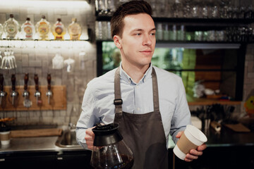 a young man of European appearance is waiting for new satisfied customers, holding a Cup of hot coffee in the hands of a paper Cup. a Barista, employee of the waiter in restaurant cafe bistro