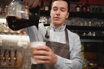a staff member in a small restaurant works in an apron and shirt. Barista waiter prepares sells coffee Americano cappuccino with a takeaway in the cafe. pours a paper Cup from the teapot into a Cup.