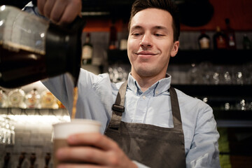 Barista waiter prepares sells coffee Americano cappuccino with a takeaway in the cafe. pours a paper Cup from the teapot into a Cup. a staff member in a small restaurant works in an apron and shirt