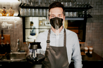 waiter male mask from the early stages of the pandemic virus on the face and antibacterial glass. holding a pot of hot coffee. It is worth waiting for new customers to work in a new small cafe.