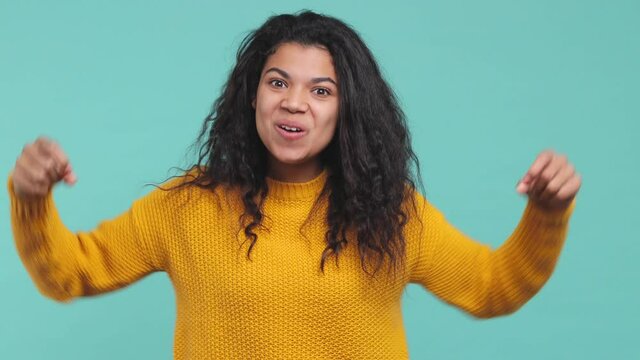 Happy young african american woman 20s in yellow sweater isolated on blue turquoise background studio. People lifestyle concept. Count countdown 1 2 3 one two three doing winner gesture point camera