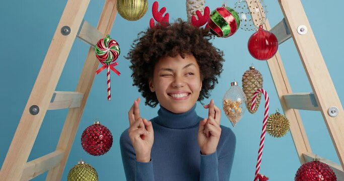 Waiting for special moment. Glad Afro American woman crosses fingers and believes in miracle happen on New Year closes eyes has overjoyed expression wears christmas reindeer antlers poses indoor