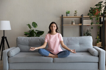 Fototapeta na wymiar Full length peaceful young arab indian multiracial woman relaxing in lotus pose on comfortable couch, enjoying practicing yoga breathing exercises alone at home, reducing stress, healthcare concept.