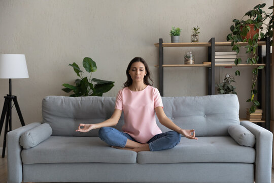Full length peaceful young arab indian multiracial woman relaxing in lotus pose on comfortable couch, enjoying practicing yoga breathing exercises alone at home, reducing stress, healthcare concept.