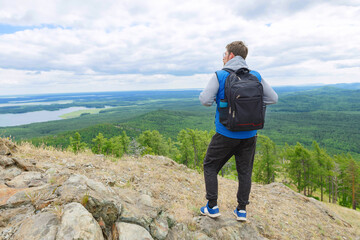 A man in a blue tank top and gray turtleneck stands on a mountain top with a full-length backpack.