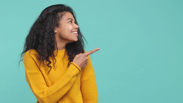 Shocked amazed young african american woman 20s years old in casual yellow sweater posing isolated on blue turquoise background in studio. People lifestyle concept. Pointing index finger aside say wow