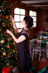 A beautiful girl in a black evening dress dresses up and decorates the Christmas tree. New Years Eve. Festive home atmosphere. Portrait of a woman. Cozy background with lights of garlands.