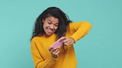 Excited funny young african american woman 20s years old in yellow sweater posing isolated on blue turquoise background studio. People lifestyle concept. Play game on mobile phone doing winner gesture