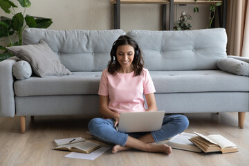 Full length happy beautiful millennial mixed race woman wearing headphones with mic, involved in video call meeting with teacher, studying remotely on online courses, sitting on warm floor at home.