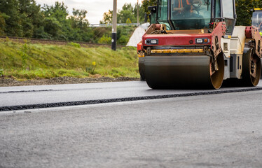Fototapeta premium Asphalt road roller with heavy vibration roller compactor press new hot asphalt on the roadway on a road construction site. Heavy Vibration roller at asphalt pavement working. Repairing.