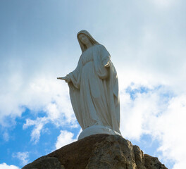 White marble statue of Notre Dame de la Serra standing on a rock against dramatic sky . Near Calvi...