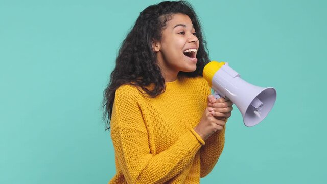 Excited young african american woman 20s in yellow sweater posing isolated on blue turquoise background studio. People lifestyle concept. Looking aside screaming in megaphone showing thumb up blinking